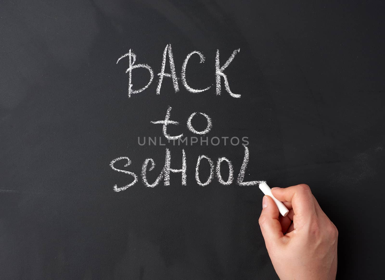 white chalk back to school on black chalkboard, hand holds a piece of white chalk, close up