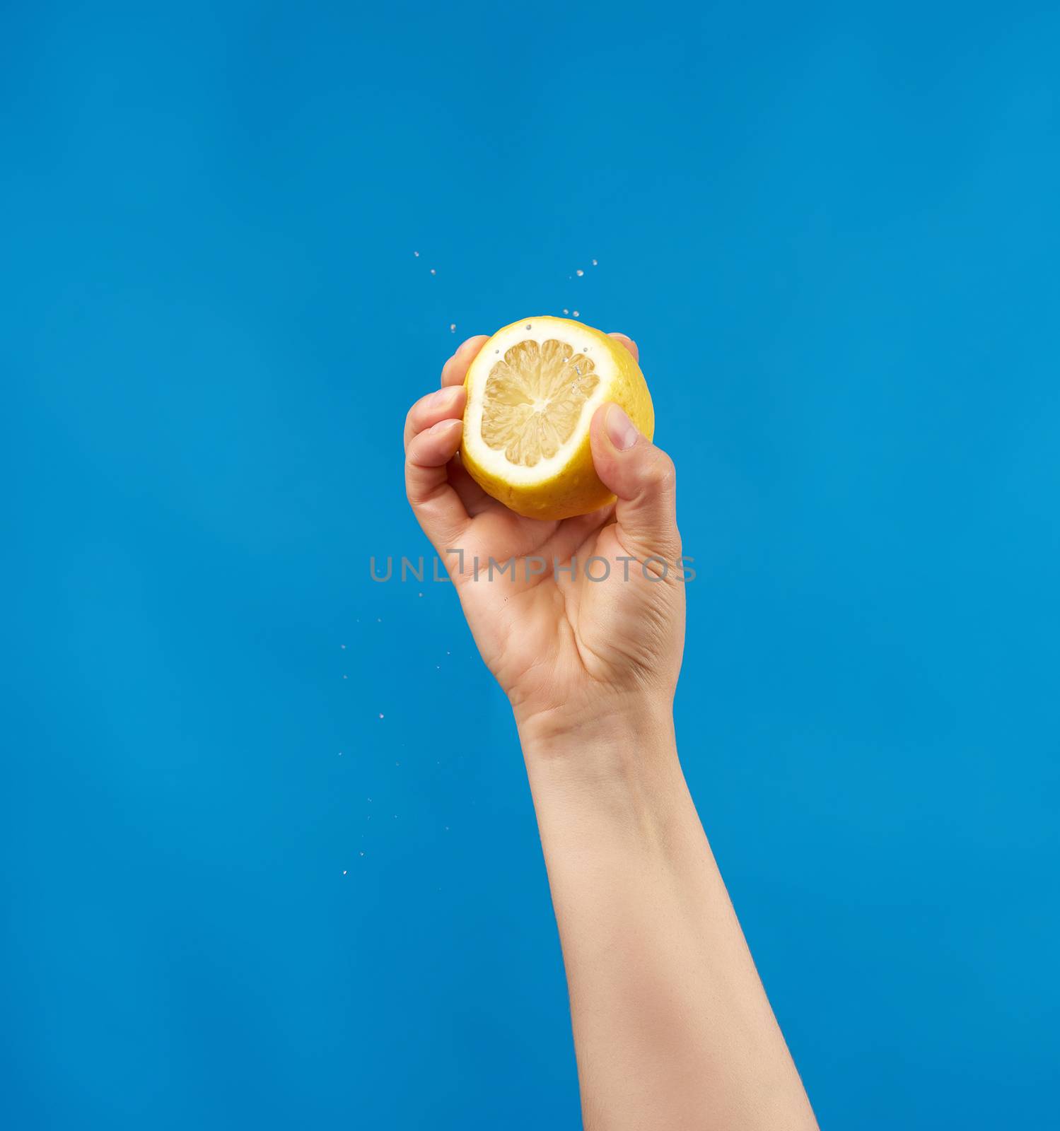 female hand holds half a yellow lemon and squeezes it on a blue background, splashes fly to the sides