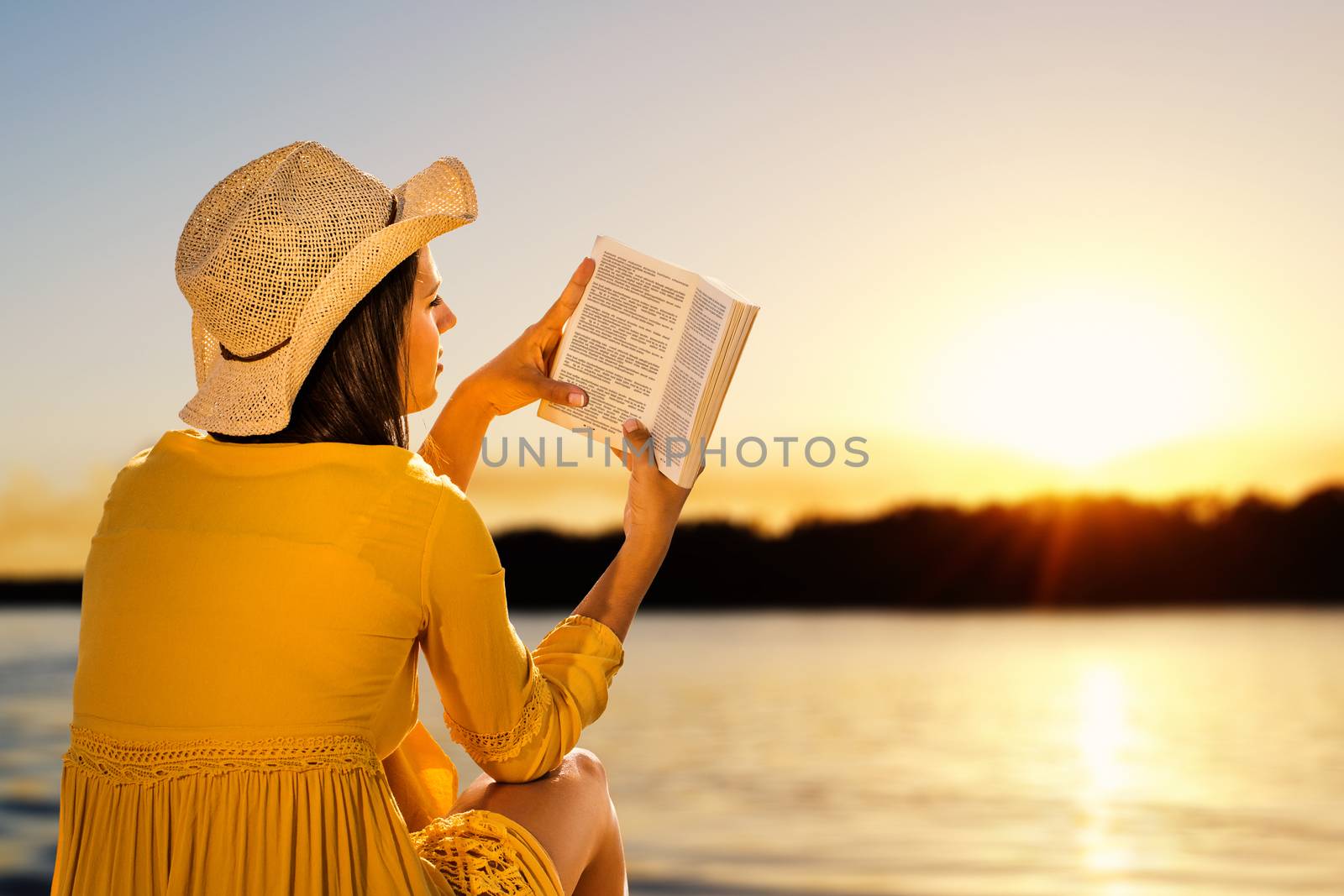 Young woman wearing hat next to river reading a book. Side view of Girl in yellow dress against sunset background.