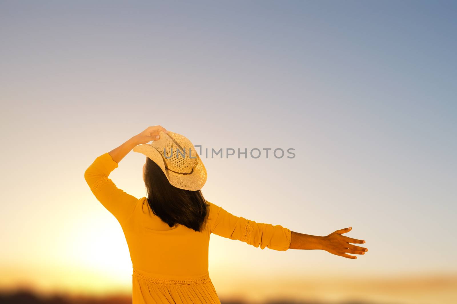 Close up rear view of young woman in yellow dress looking at sunset. Girl holding hat on head with one hand and raising other hand in sky.