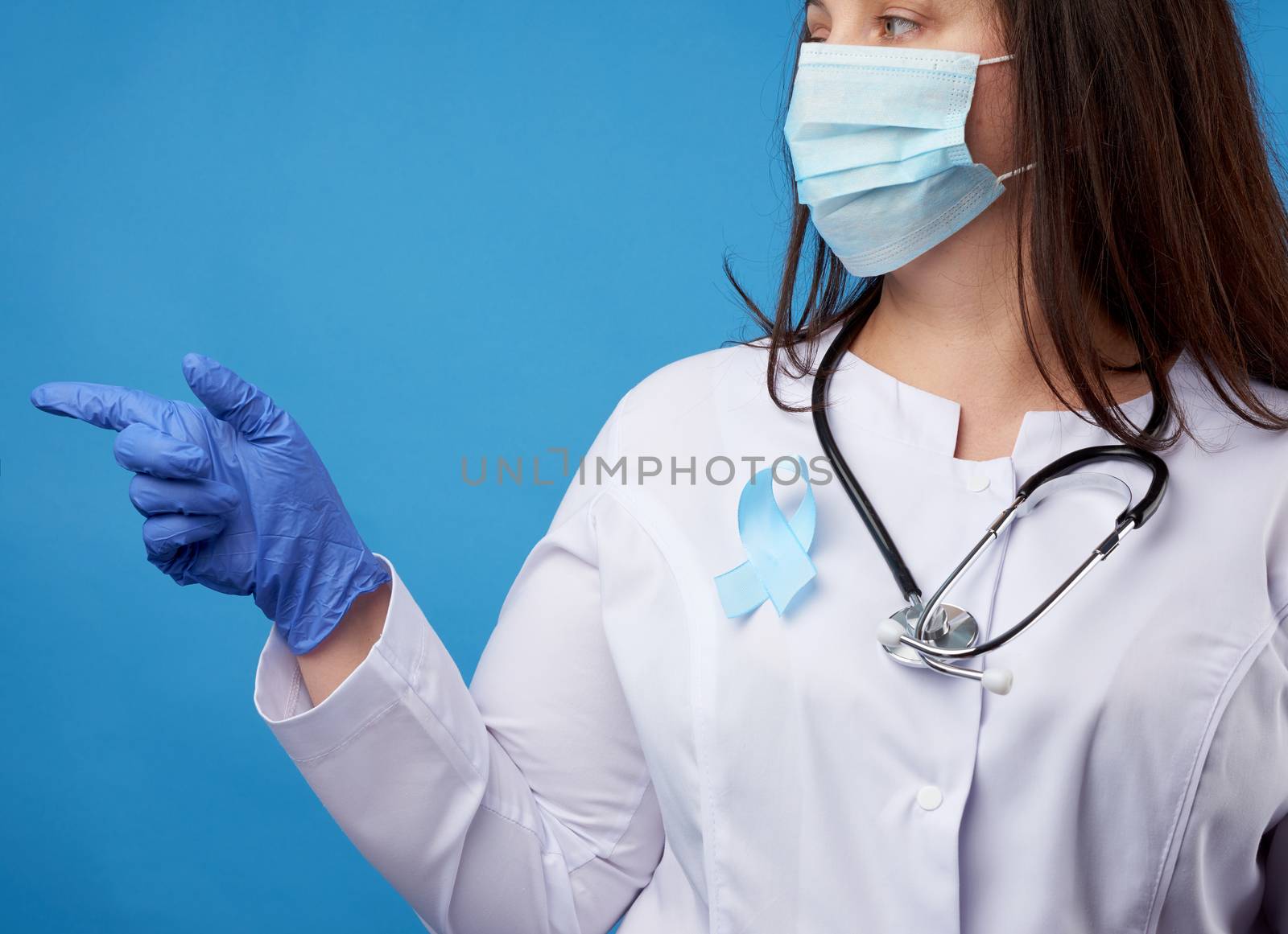 female doctor in a disposable sterile mask and a white coat stands on a blue background and points with the index finger to the side, a blue silk ribbon in the shape of a loop pinned to the chest