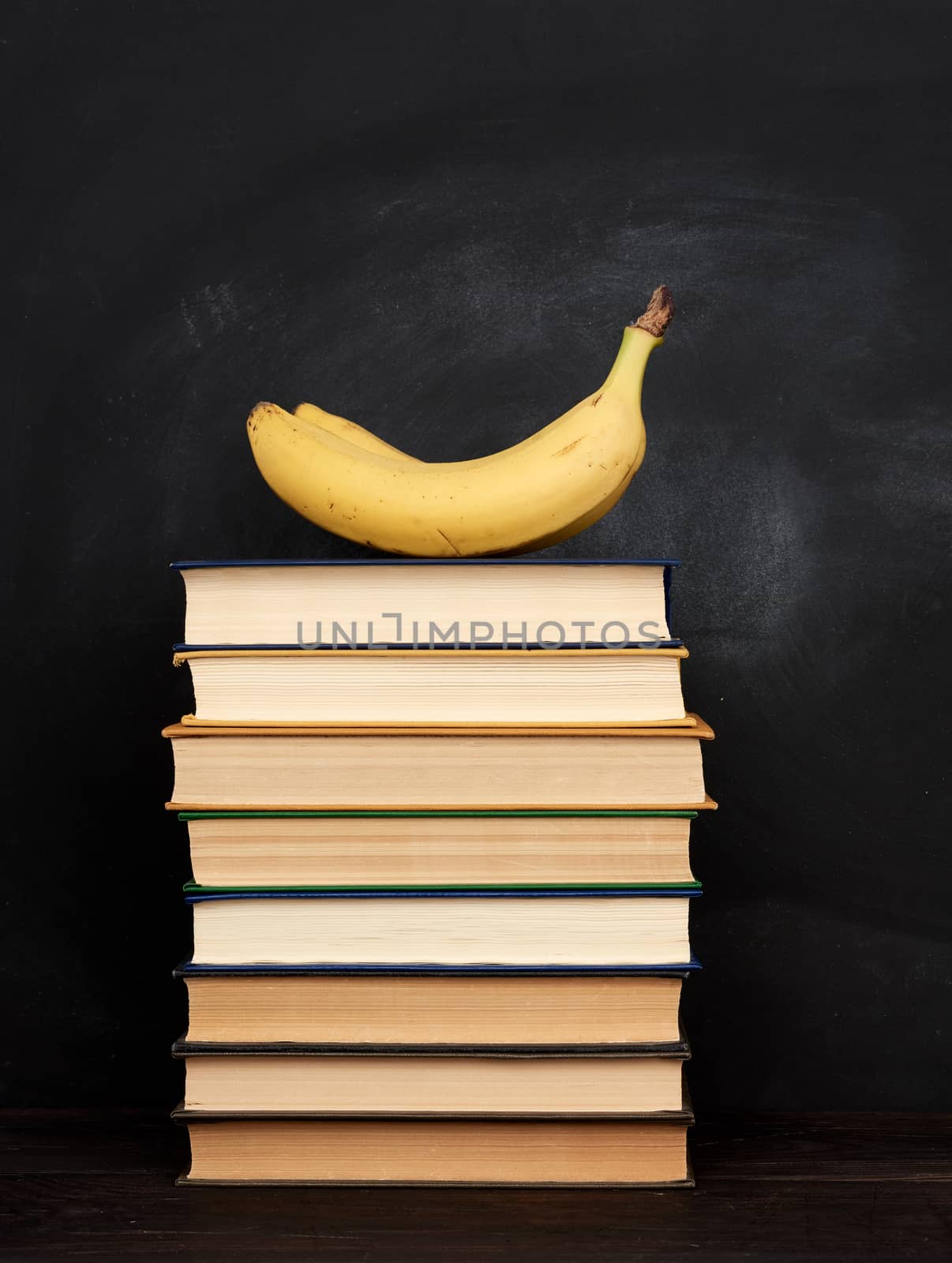 yellow ripe banana and stack of various hardback books on the background of an empty black chalk board, back to school