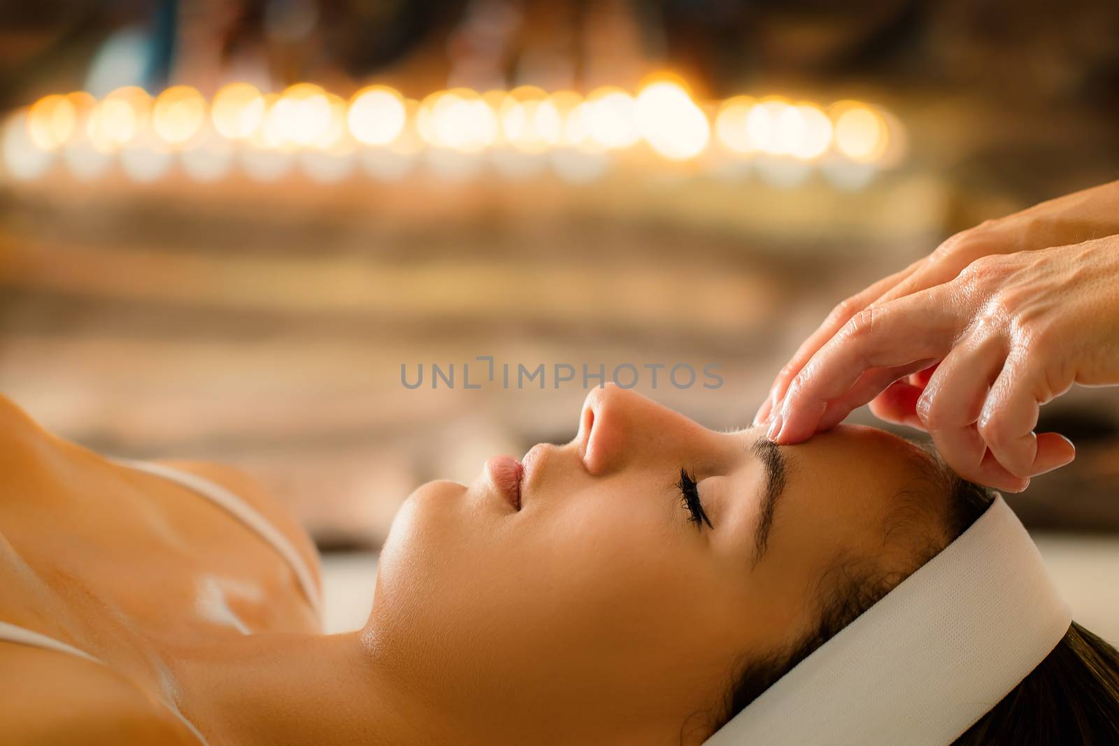 Close up portrait of woman having head massage in spa. Low key atmosphere with out of focus candles glowing in background. Therapist touching girl’s forehead.