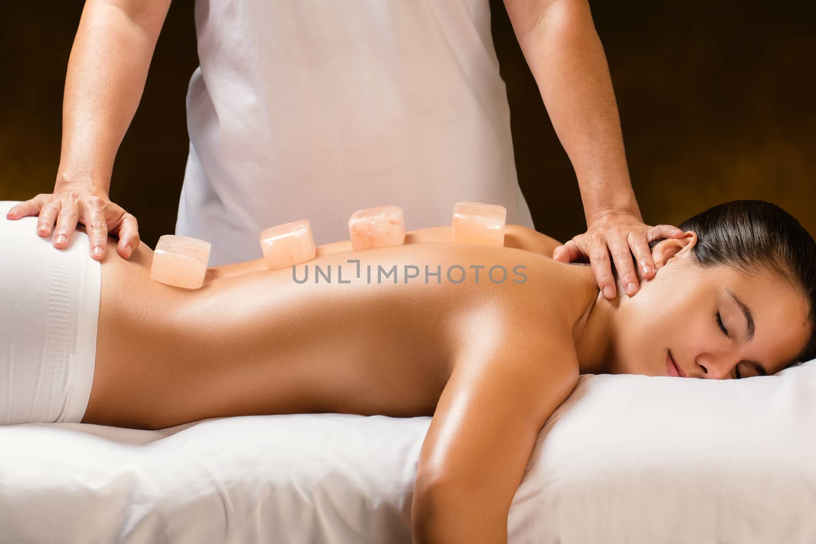 Close up of young woman having hot himalayan stone massage in spa.Natural salt stone bricks placed on spine with therapist in background.