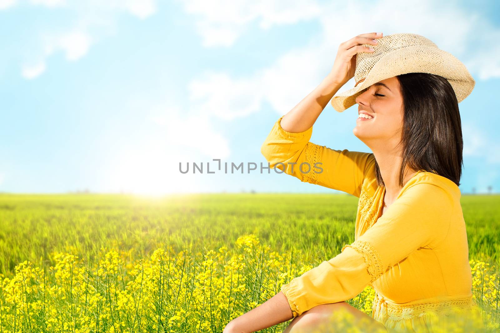 Close up portrait of attractive young woman in yellow dress sitting in green flower field.Happy girl wearing hat smiling with eyes closed enjoying nature.