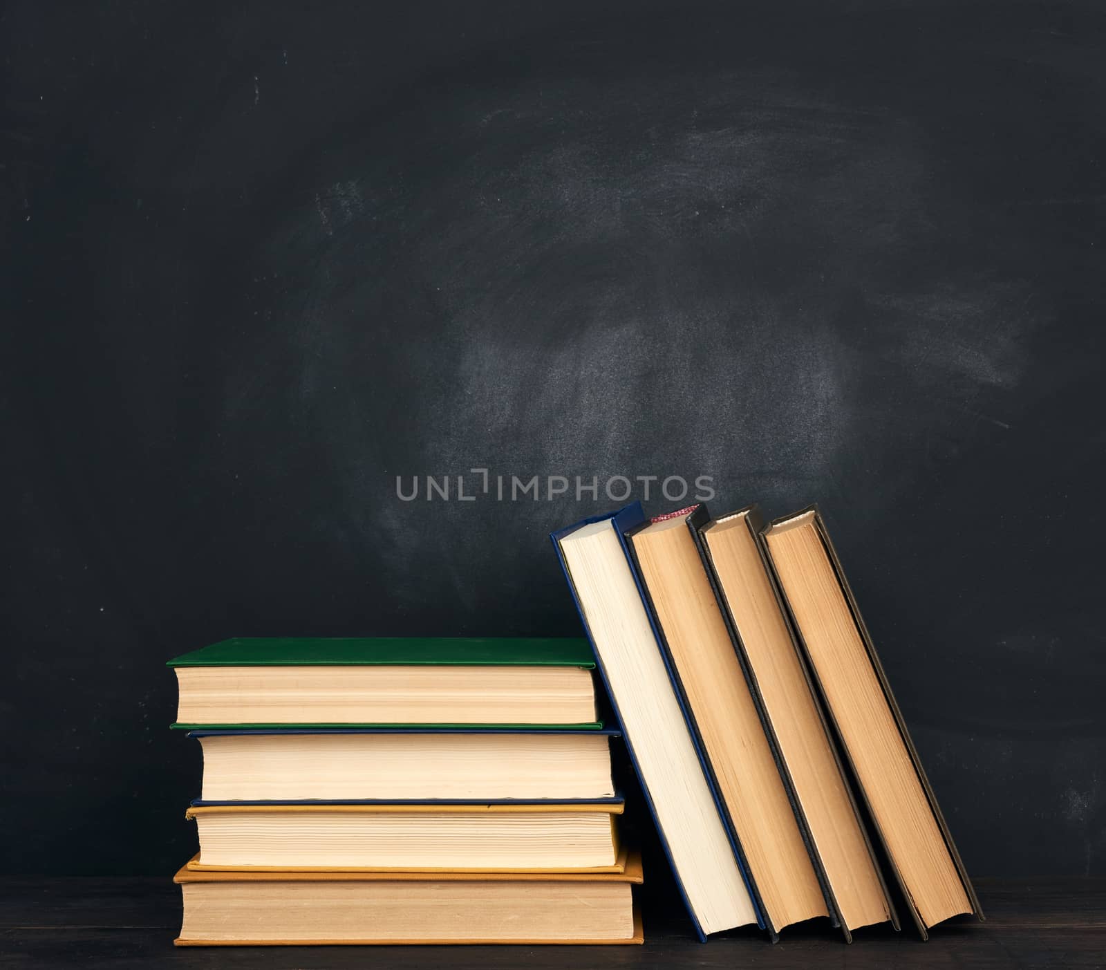 stack of various hardback books on the background of an empty black chalk board, place for text, back to school