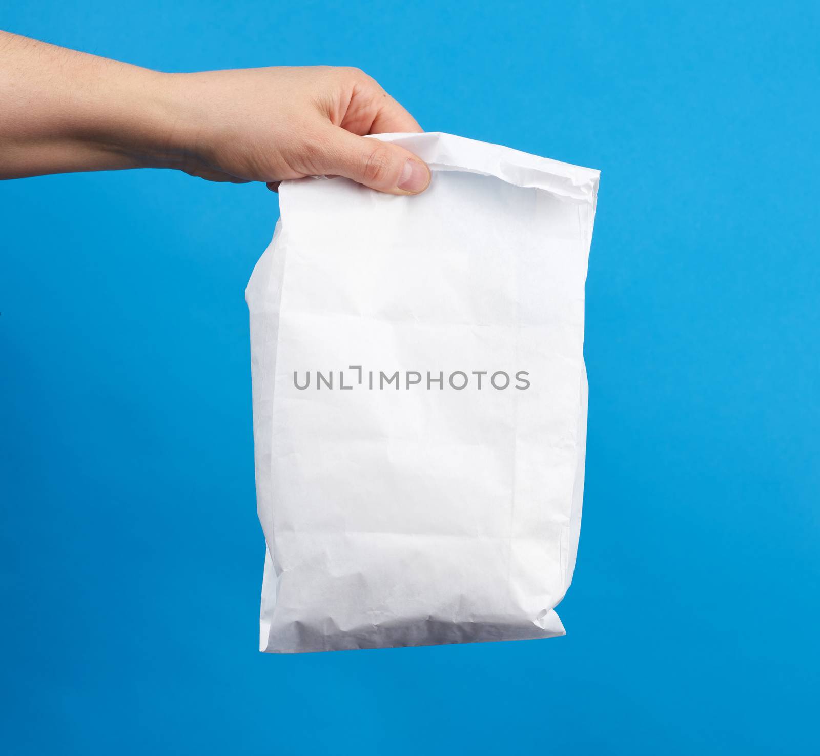 female hand holds a disposable full white paper bag, takeaway and delivery food concept, blue background