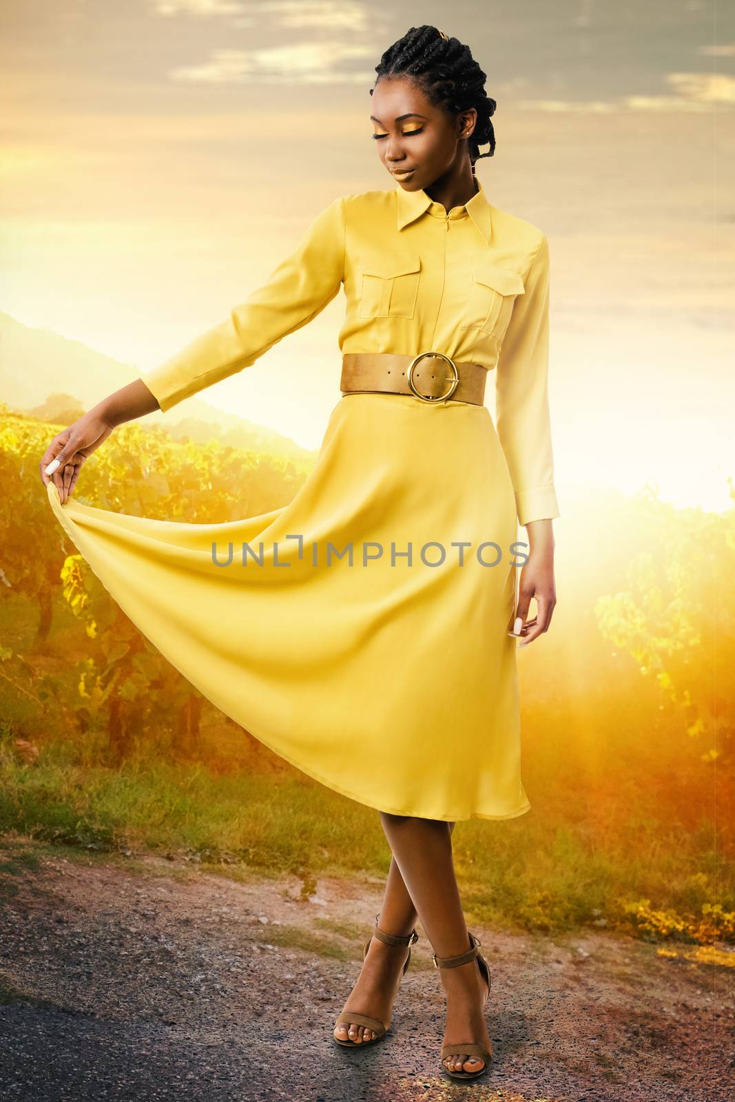 Full length portrait of attractive african american girl standing in vineyard at sunset. Young woman in stylish yellow dress with eyes closed.