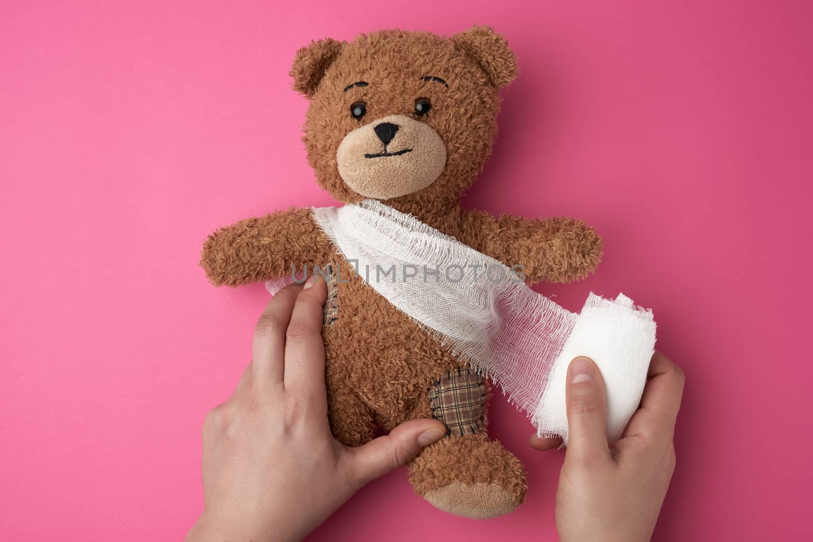brown teddy bear with bandaged torso with white gauze bandage on a pink background, two female hands are holding a toy