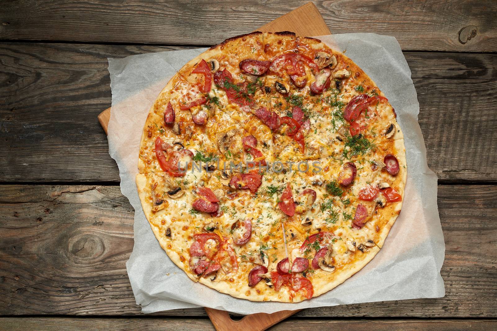 baked round pizza with smoked sausages, mushrooms, tomatoes, cheese, food is cut in portions, wooden table 