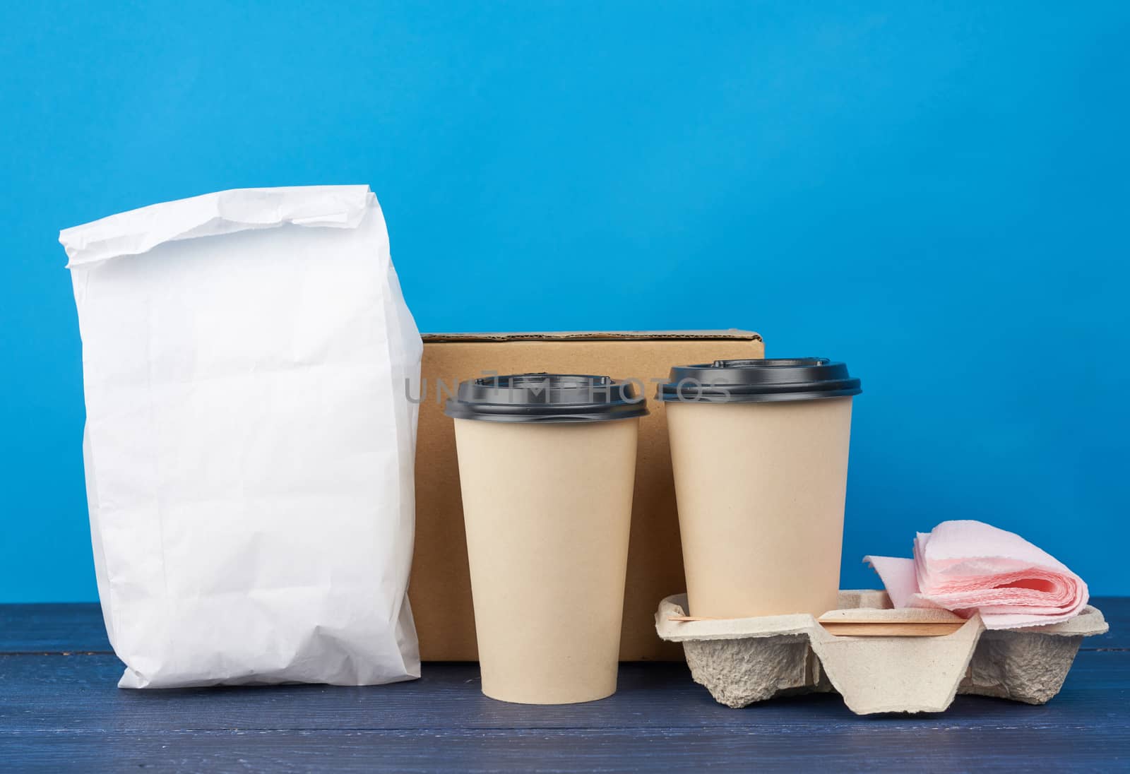 full white paper bag and brown paper disposable cups with a plastic lid stand in the tray on a blue wooden table, takeaway containers
