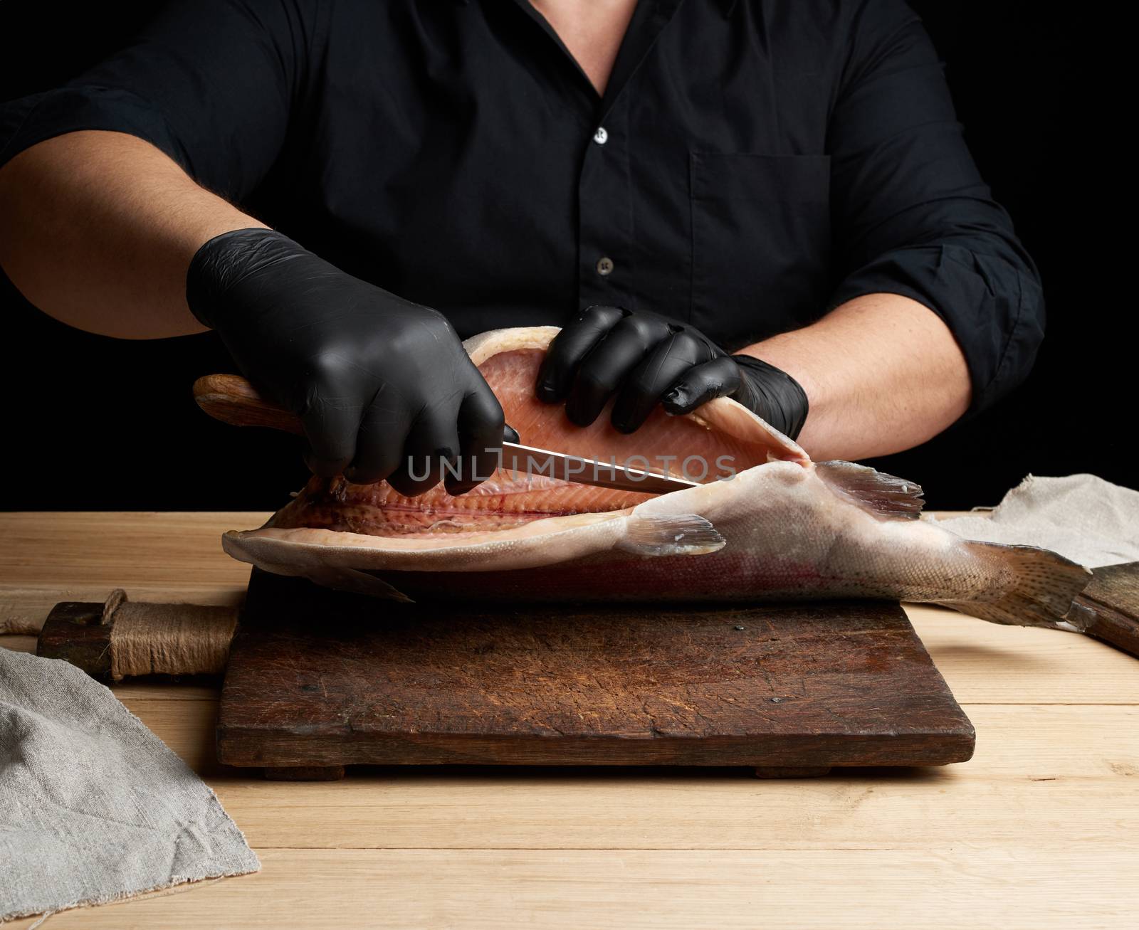chef in black uniform and black latex gloves cuts a carcass of fresh salmon fish on a wooden cutting board, black background