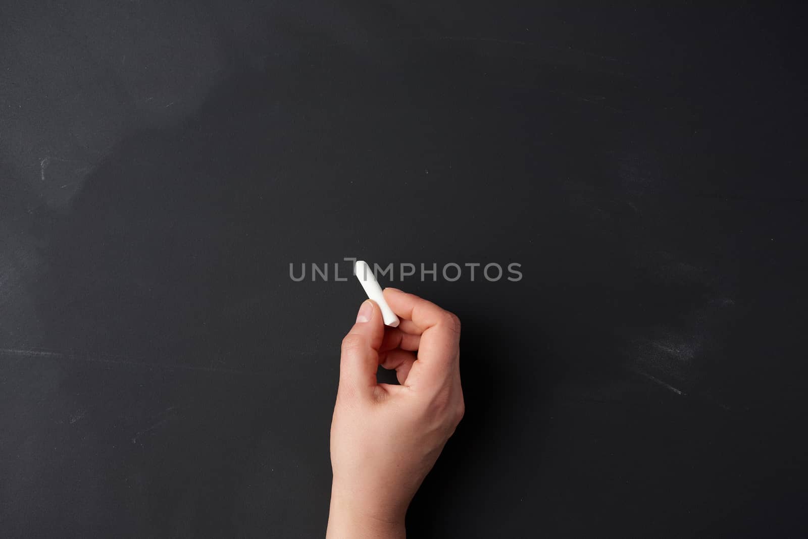 hand holds a piece of white chalk on the background of an empty black chalk board, presentation concept, back to school