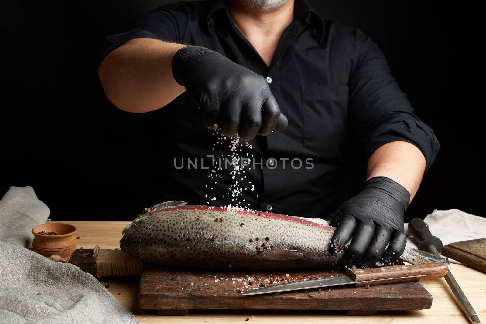 chef in a black shirt and black latex gloves prepares salmon fillet on a wooden cutting board, process of sprinkling with spices and salt, low key