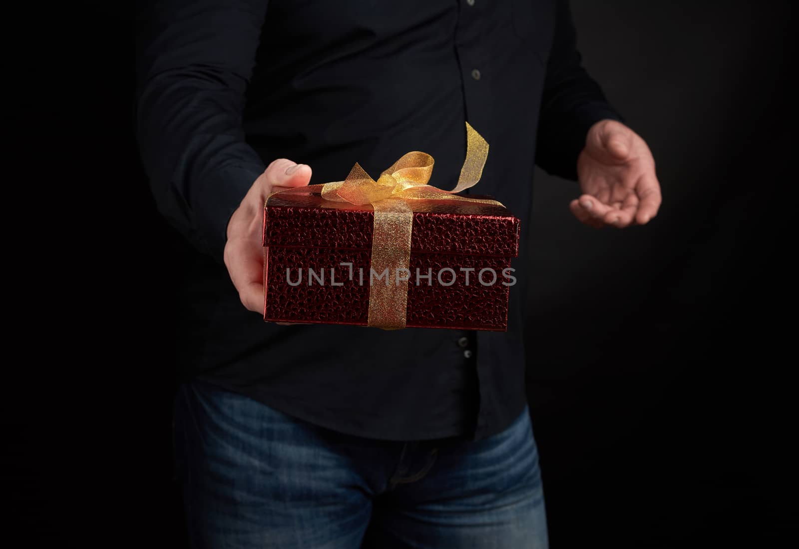 adult man in a black shirt holds a red square box with a knotted golden bow on a dark background, concept of congratulations, surprise