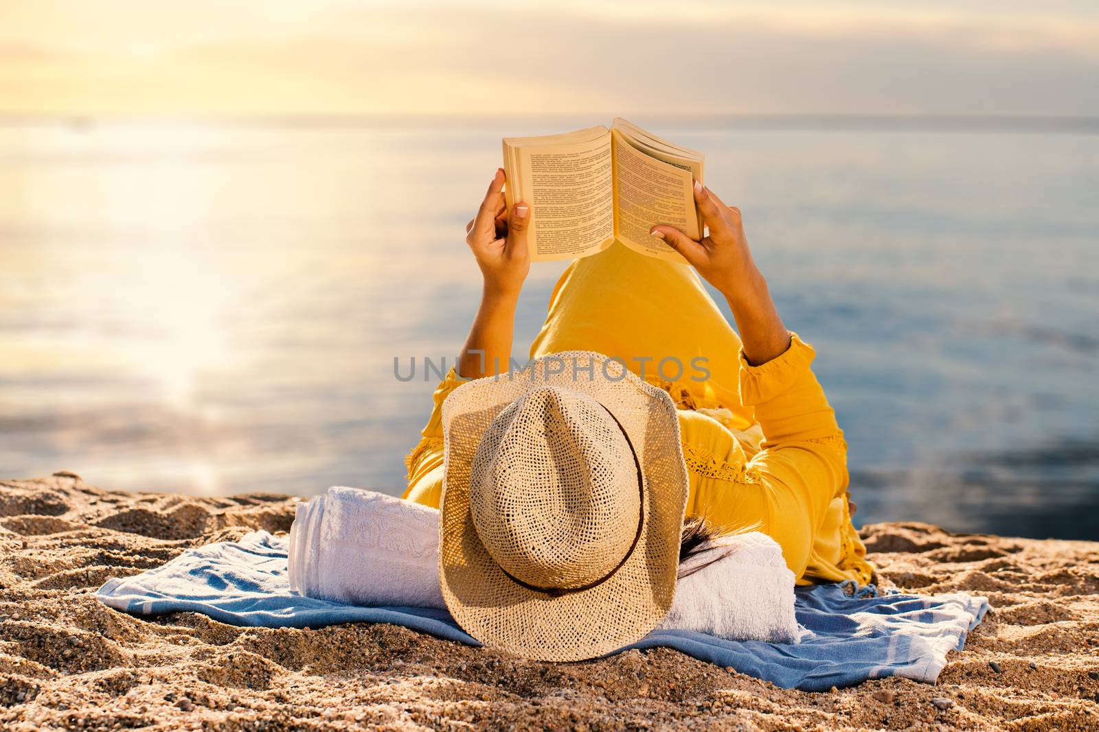 Close up of woman in yellow dress laying on beach reading a book. Rear view of woman wearing hat against idyllic sunset.