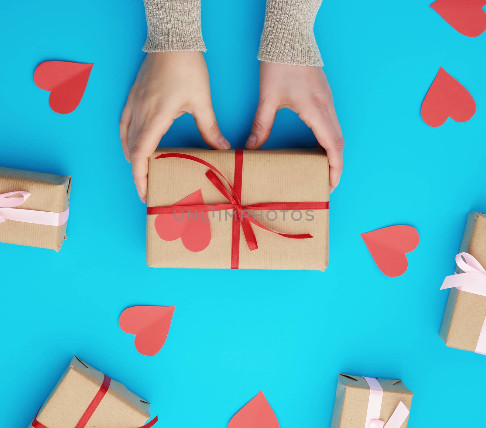 female hand holds a box wrapped in brown kraft paper and tied with a silk ribbon on a blue background, paper red hearts. Valentine's Day backdrop