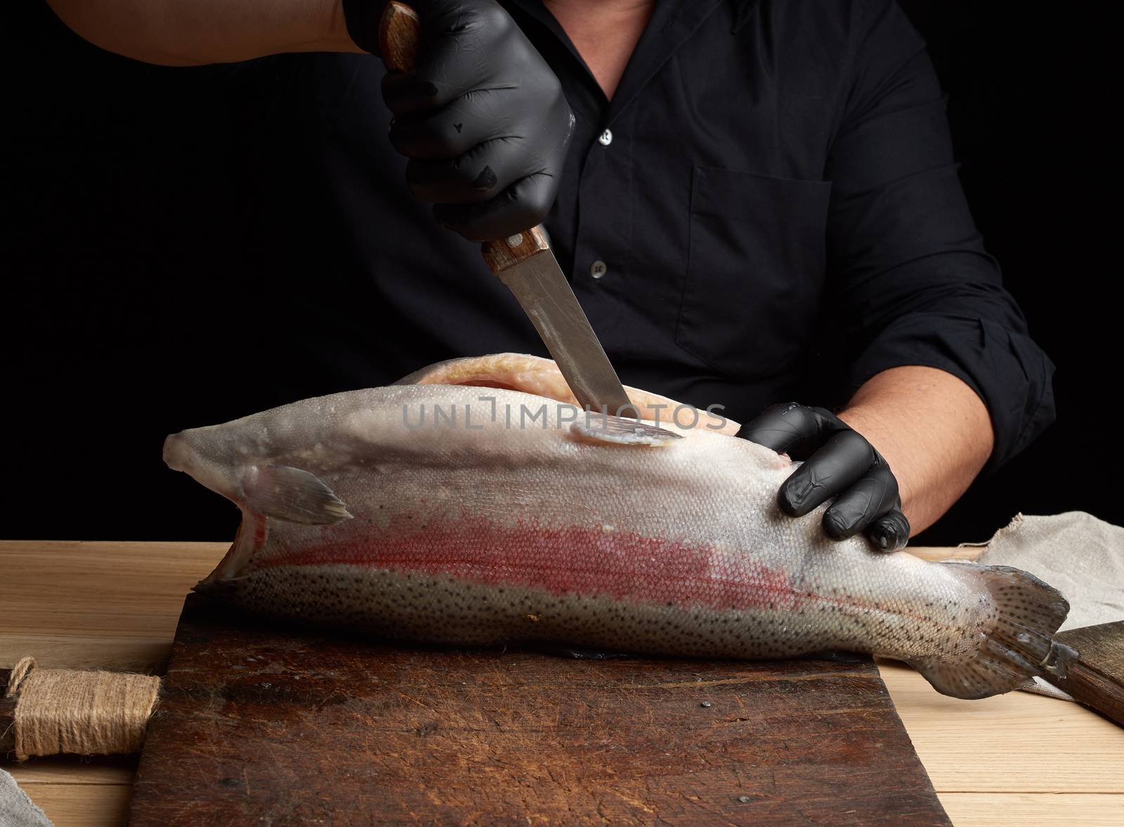 chef in black shirt and black latex gloves slices a whole fresh fish salmon on a vintage brown cutting board,  low key