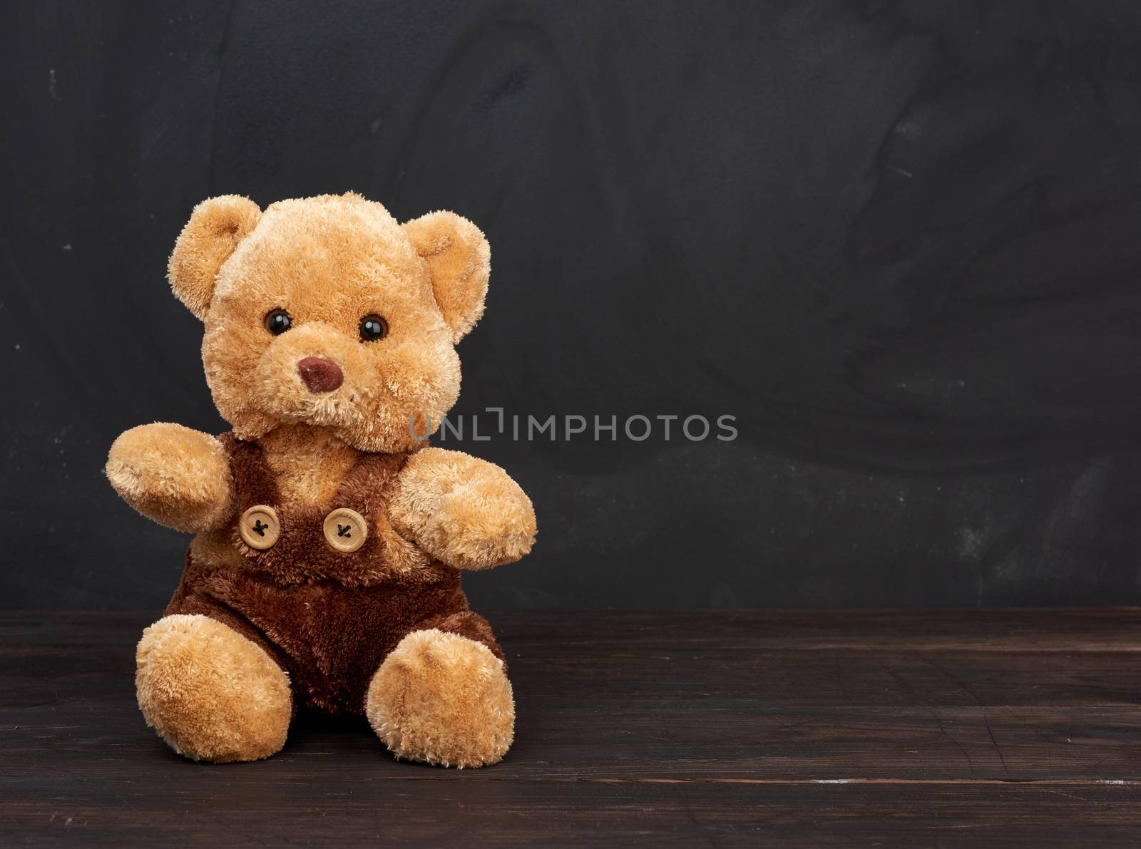 brown teddy bear sits on a brown wooden table, behind an empty black chalk board, place for an inscription, back to school