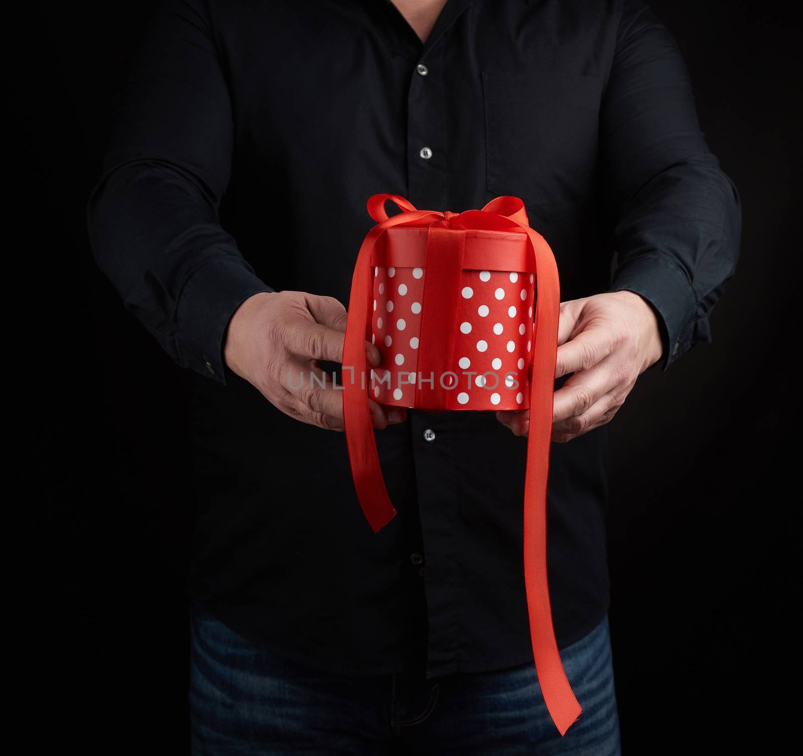 adult man in a black shirt holds in his hand a round red box of polka dots with a red bow on a black background, concept of a holiday greetings, surprise and gratitude
