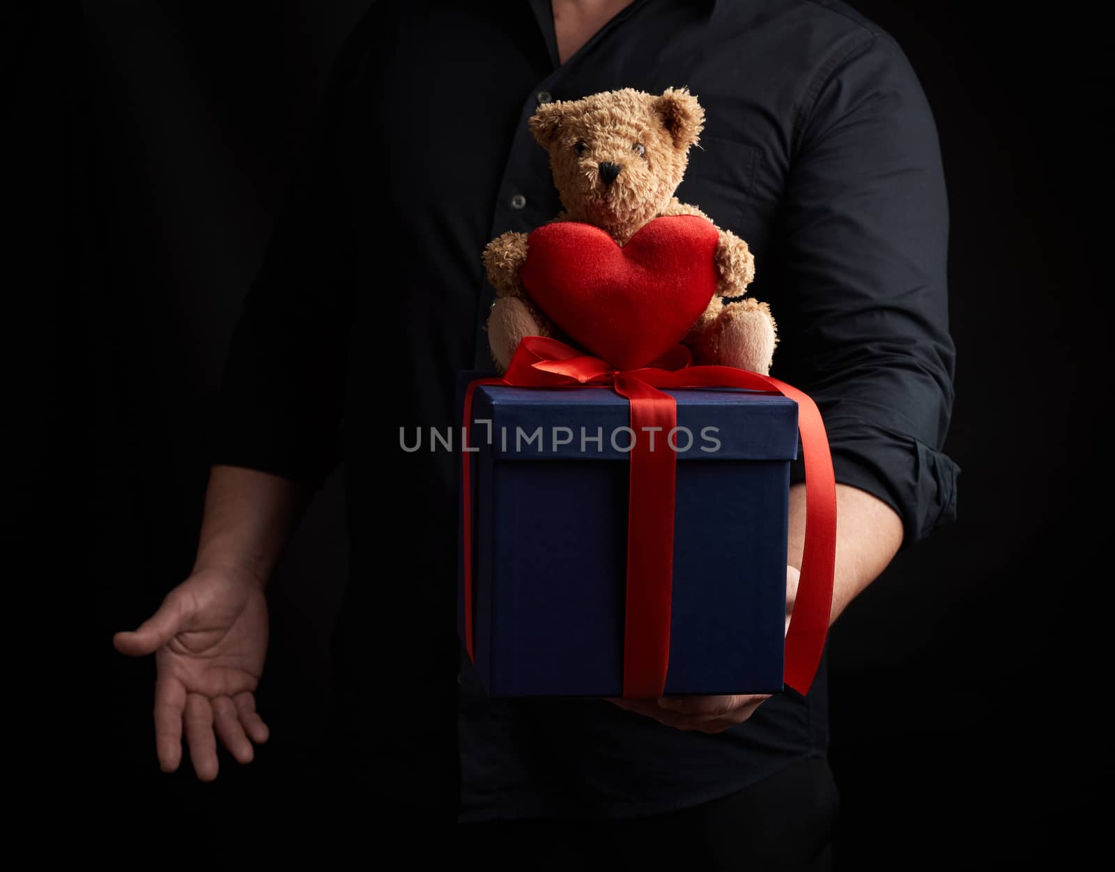 adult man in a black shirt holds a blue square box tied with a red ribbon and sits on top of a brown teddy bear with a heart, concept of congratulations on Valentine's Day on February 14