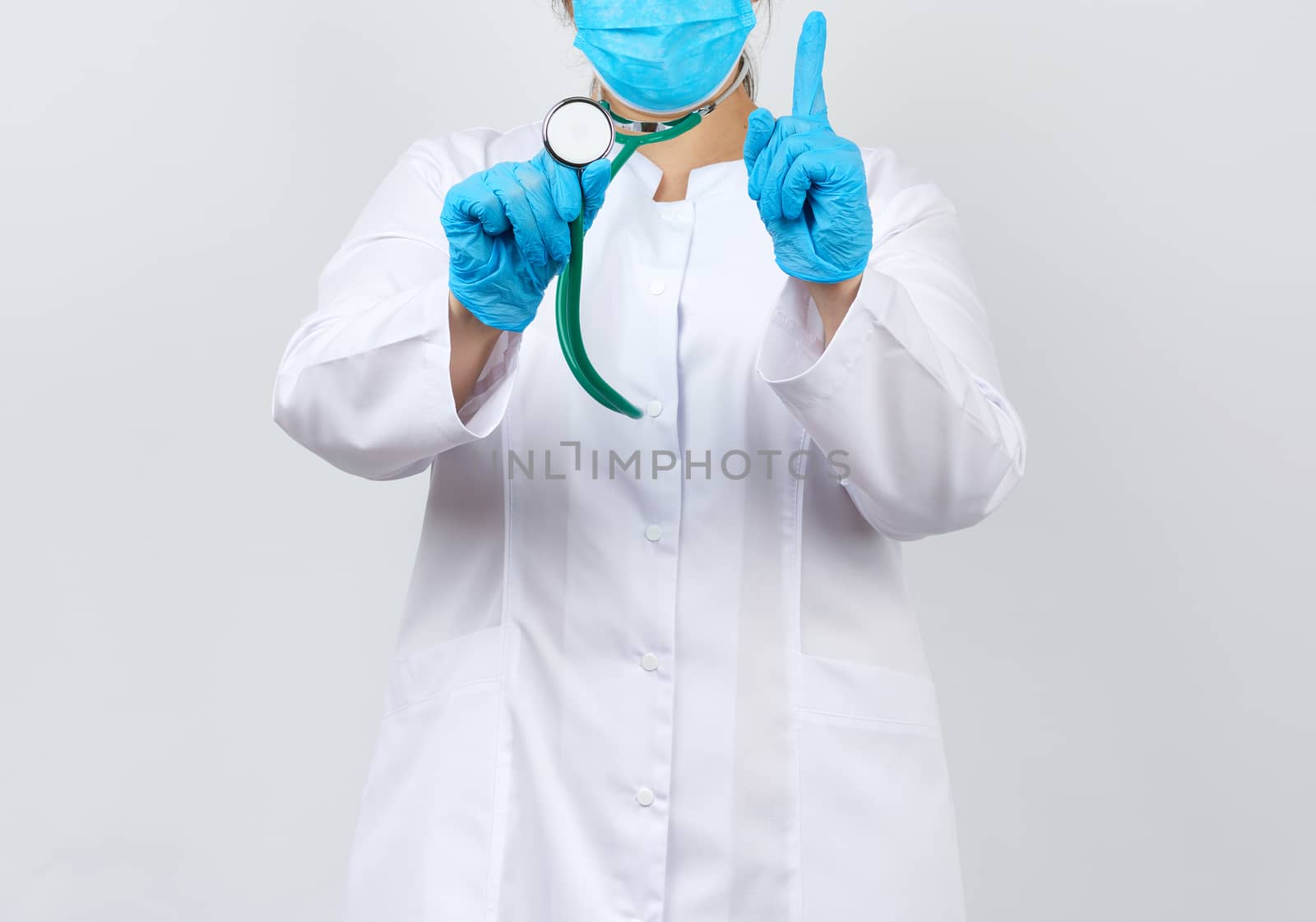 medic woman in a white coat and mask, wearing blue medical latex gloves, holds a green stethoscope on a white background, studio photo
