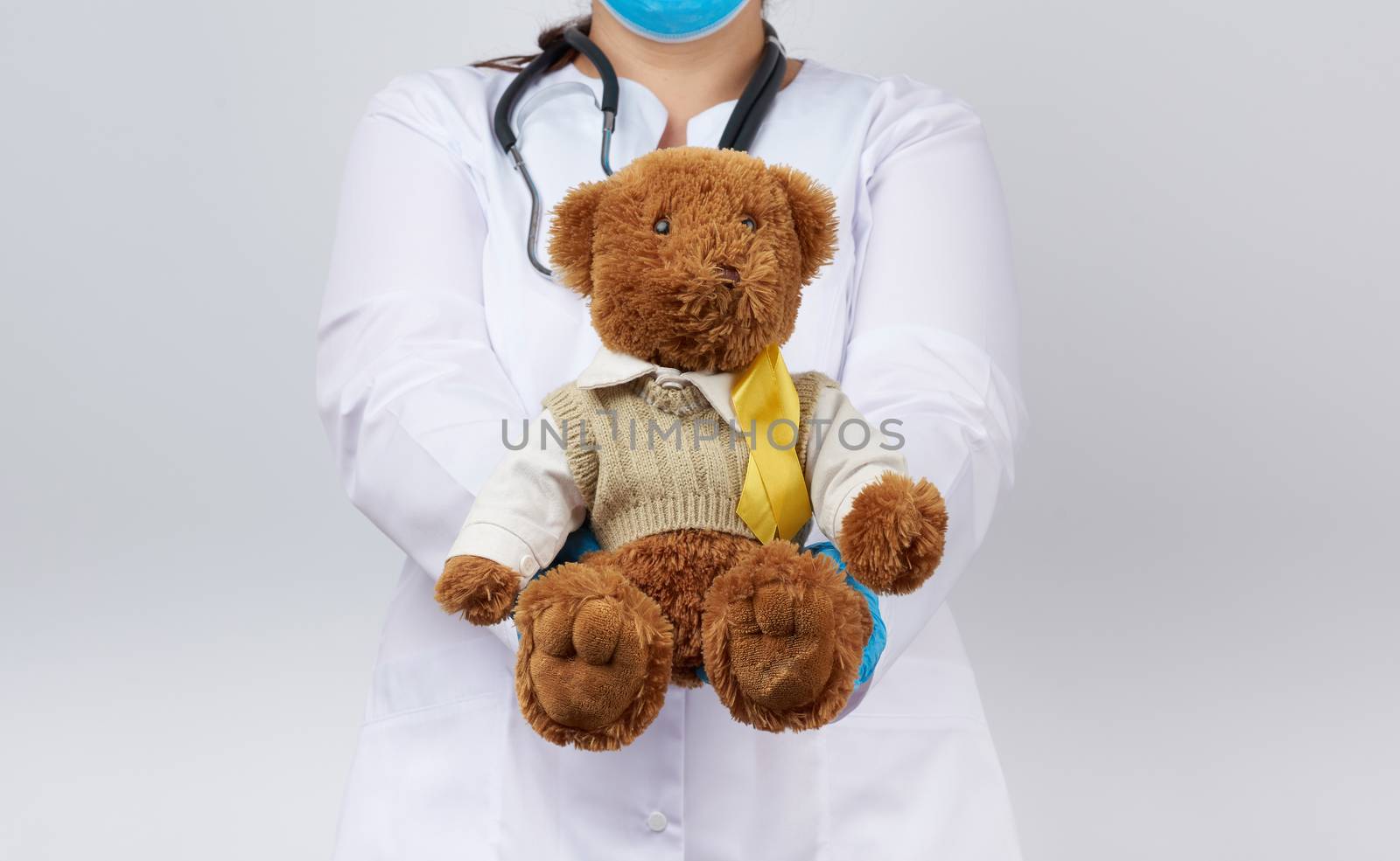 pediatrician in white coat, blue latex gloves holds a brown teddy bear with a yellow ribbon on a sweater, concept of the fight against childhood cancer, problem of suicides