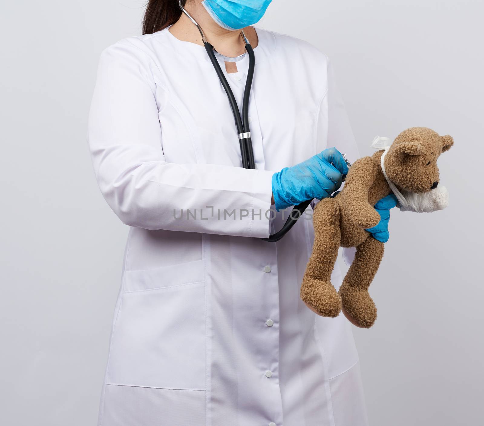 female medic holds brown teddy bear with paw bandaged in white bandage and listens to toy with stethoscope, concept of pediatrics and animal treatment