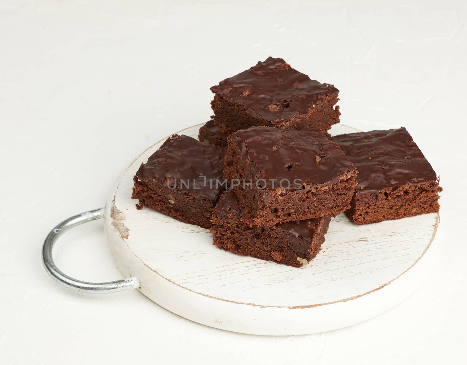 stack of square baked slices of brownie chocolate cake with walnuts on a round white wooden board, top view
