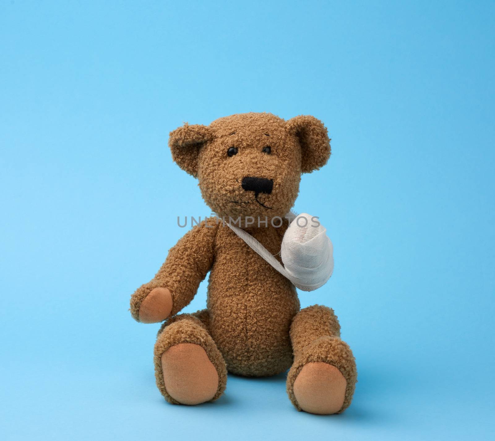 funny vintage brown curly teddy bear with rewound paw with white gauze bandage sitting on blue background, concept of injuries in children or animals