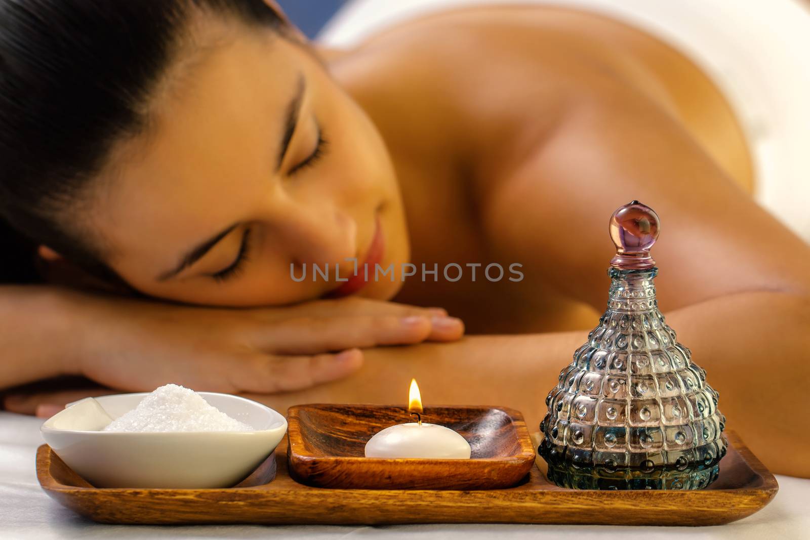 Close up still life of massage oil, candle and Himalayan salt. Out of focus woman relaxing with eyes closed in background.