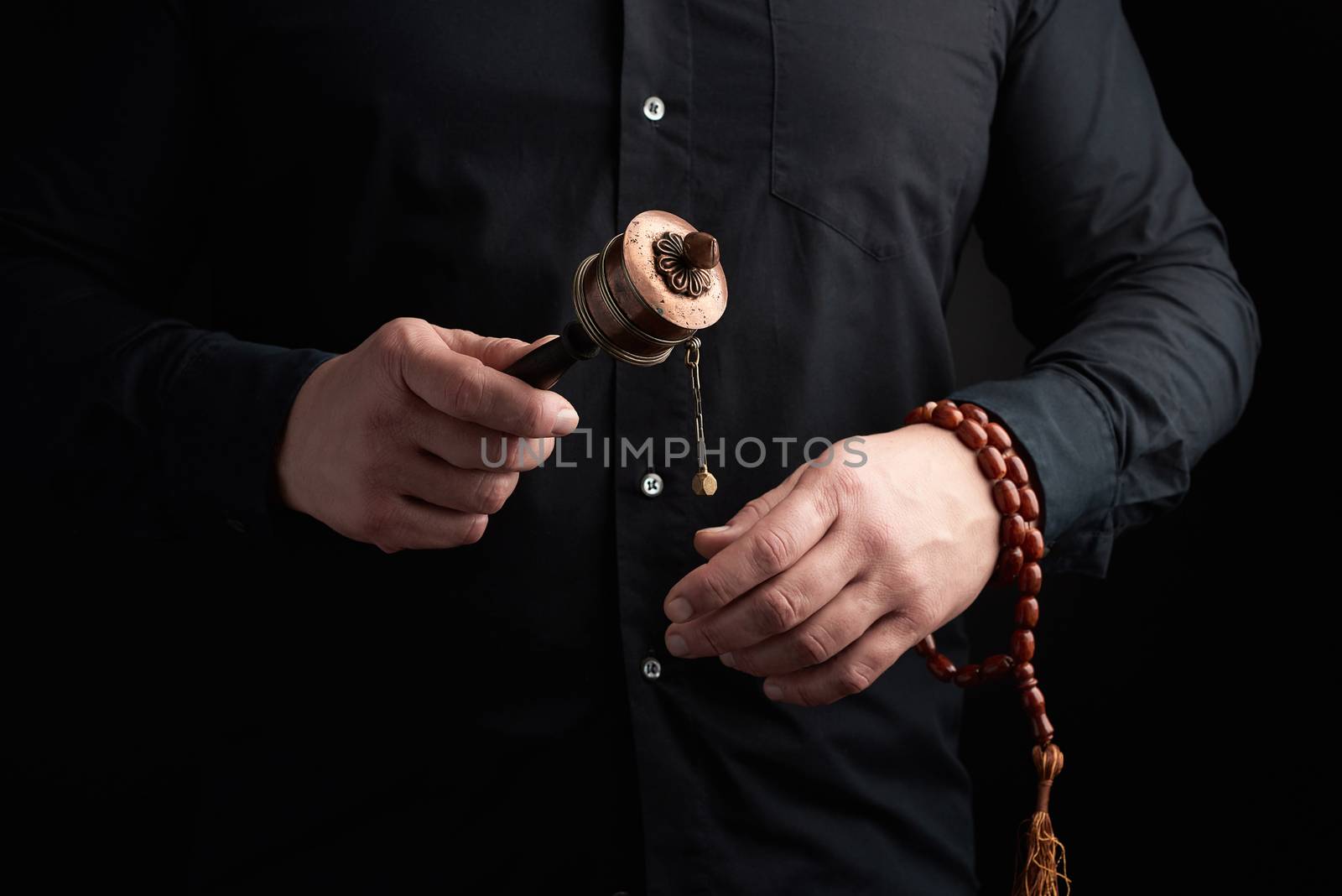 man in black clothes holds a bronze prayer drum on a wooden handle, concept of prayers and meditations