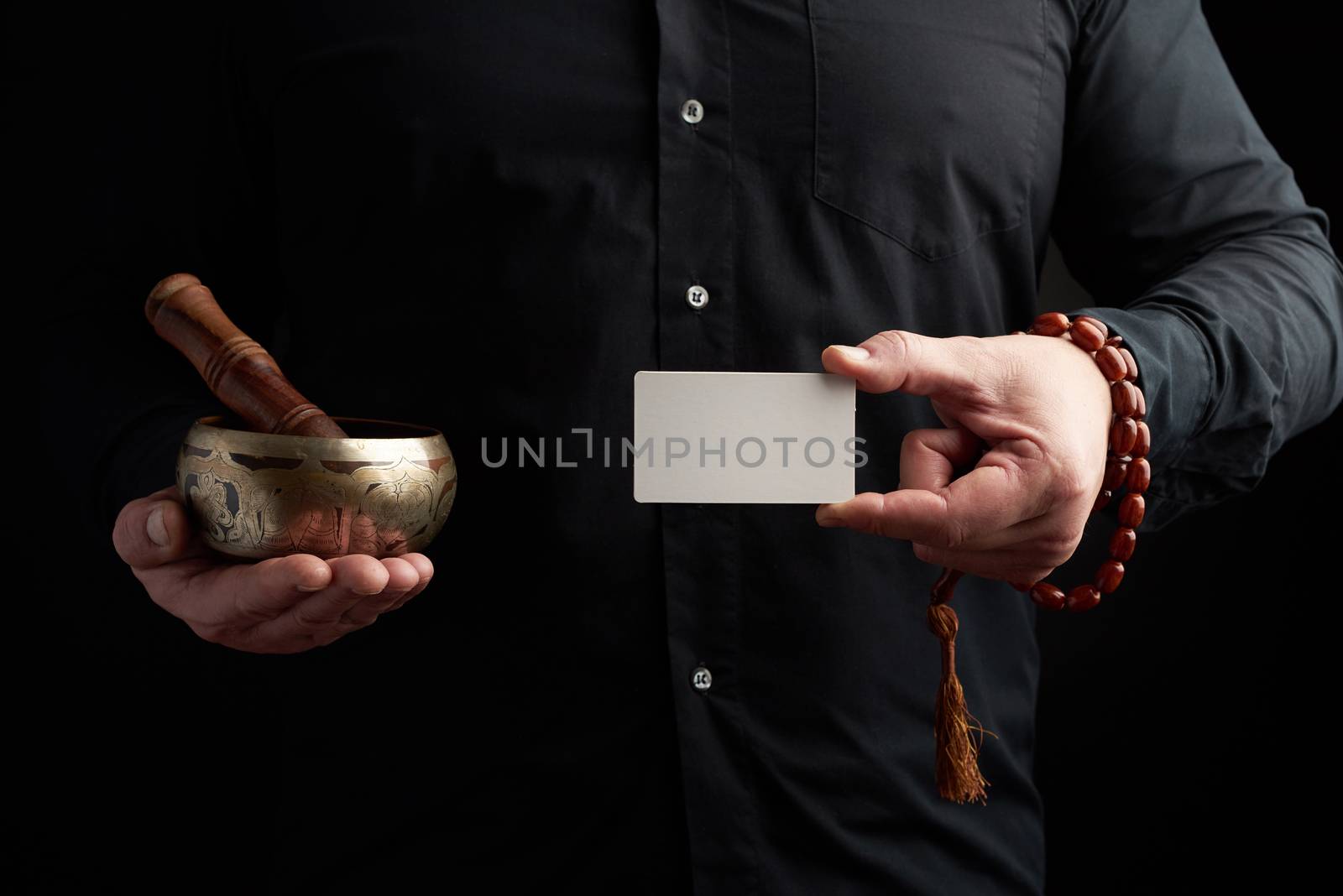 adult man in black clothes holds in his hands a copper singing bowl, object for religious rituals, meditations and alternative medicine, in his left hand is an empty white business card, a place for inscription