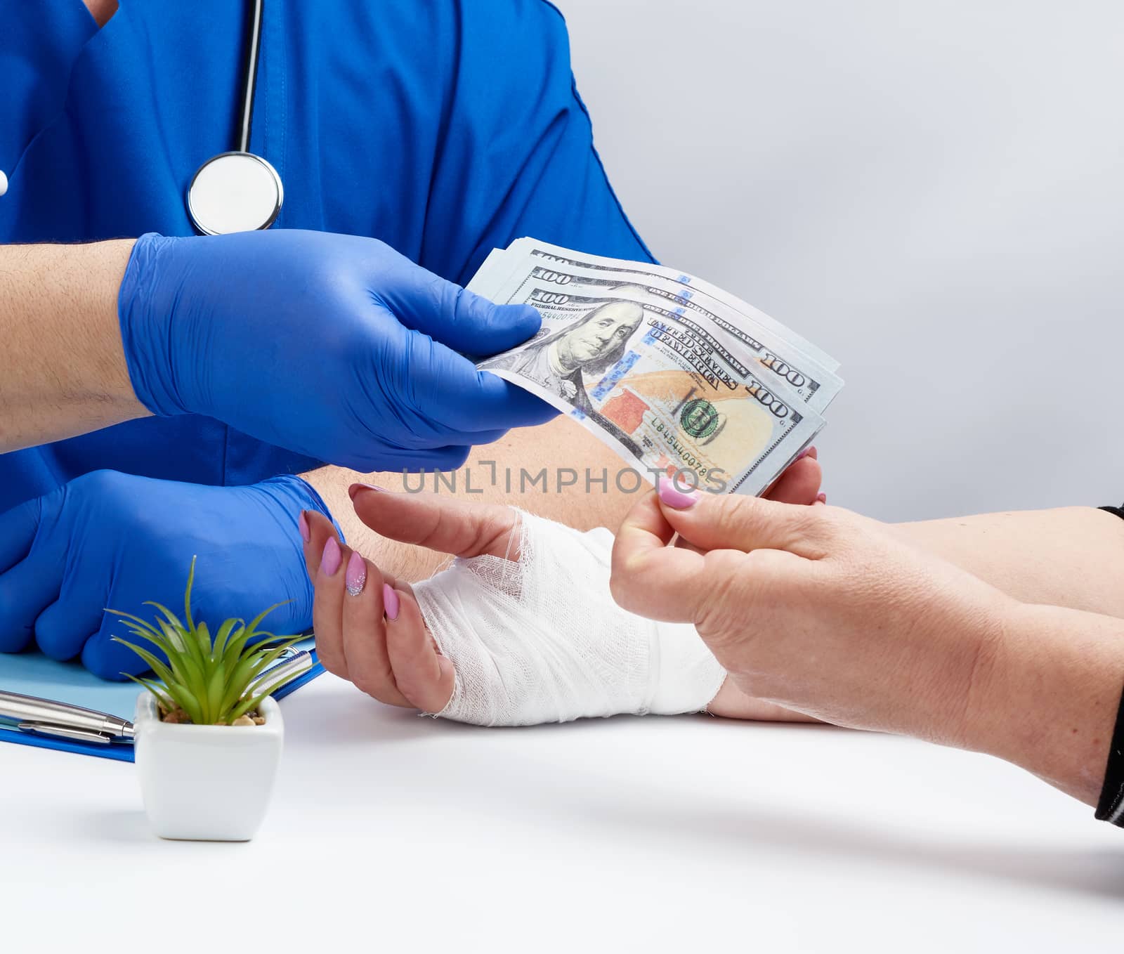 doctor in blue uniform and latex medical gloves sits at a desk and takes a wad of US dollars from a patient, concept of payment for services, cost of medical examinations