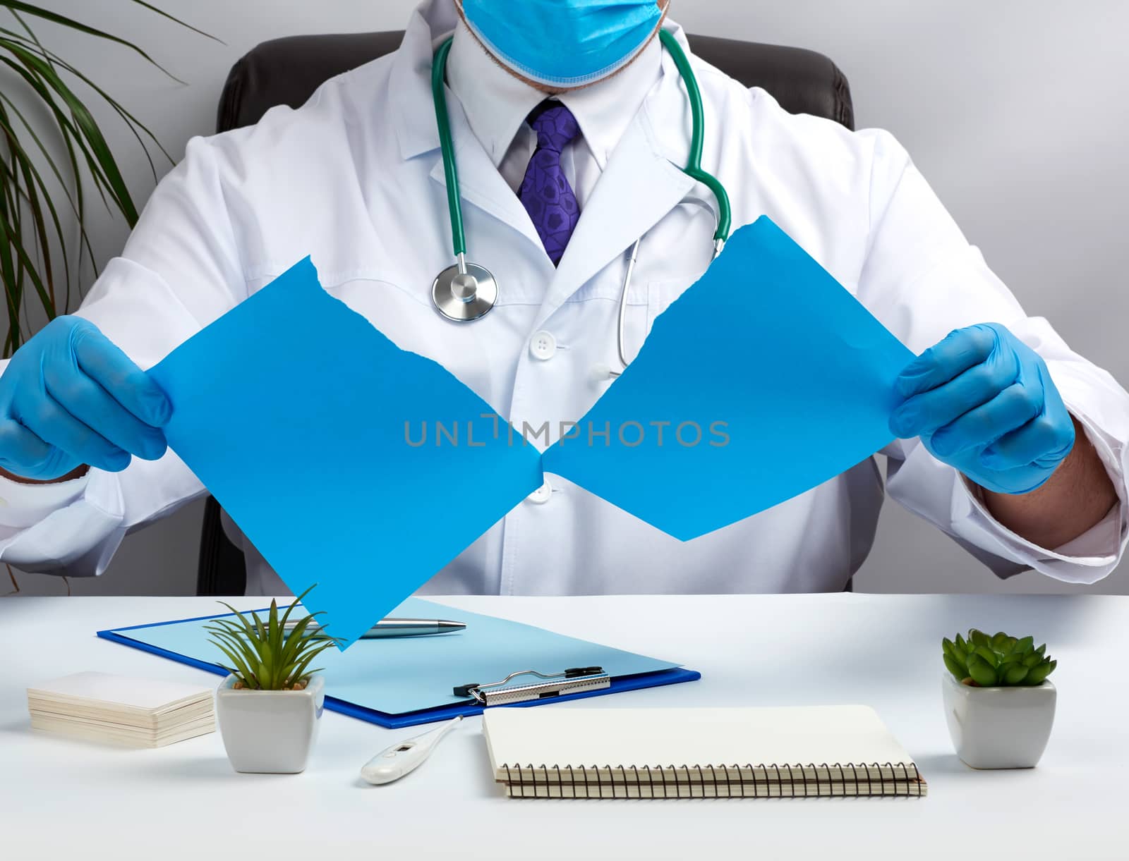 doctor in a white medical coat is sitting at a table in a brown leather chair and holding a torn blue sheet of paper in his hands, place for an inscription, a medical office