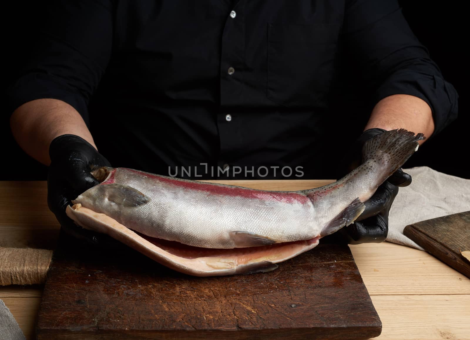 chef in a black shirt and black latex gloves holds a raw carcass of headless salmon fish over a brown wooden cutting board