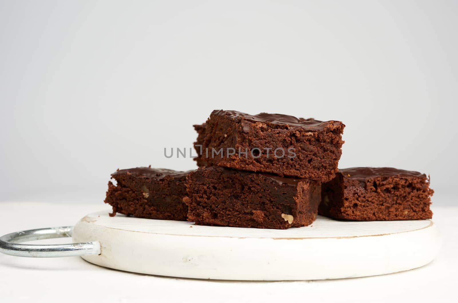stack of square baked slices of brownie chocolate cake with walnuts on a round white wooden board, close up