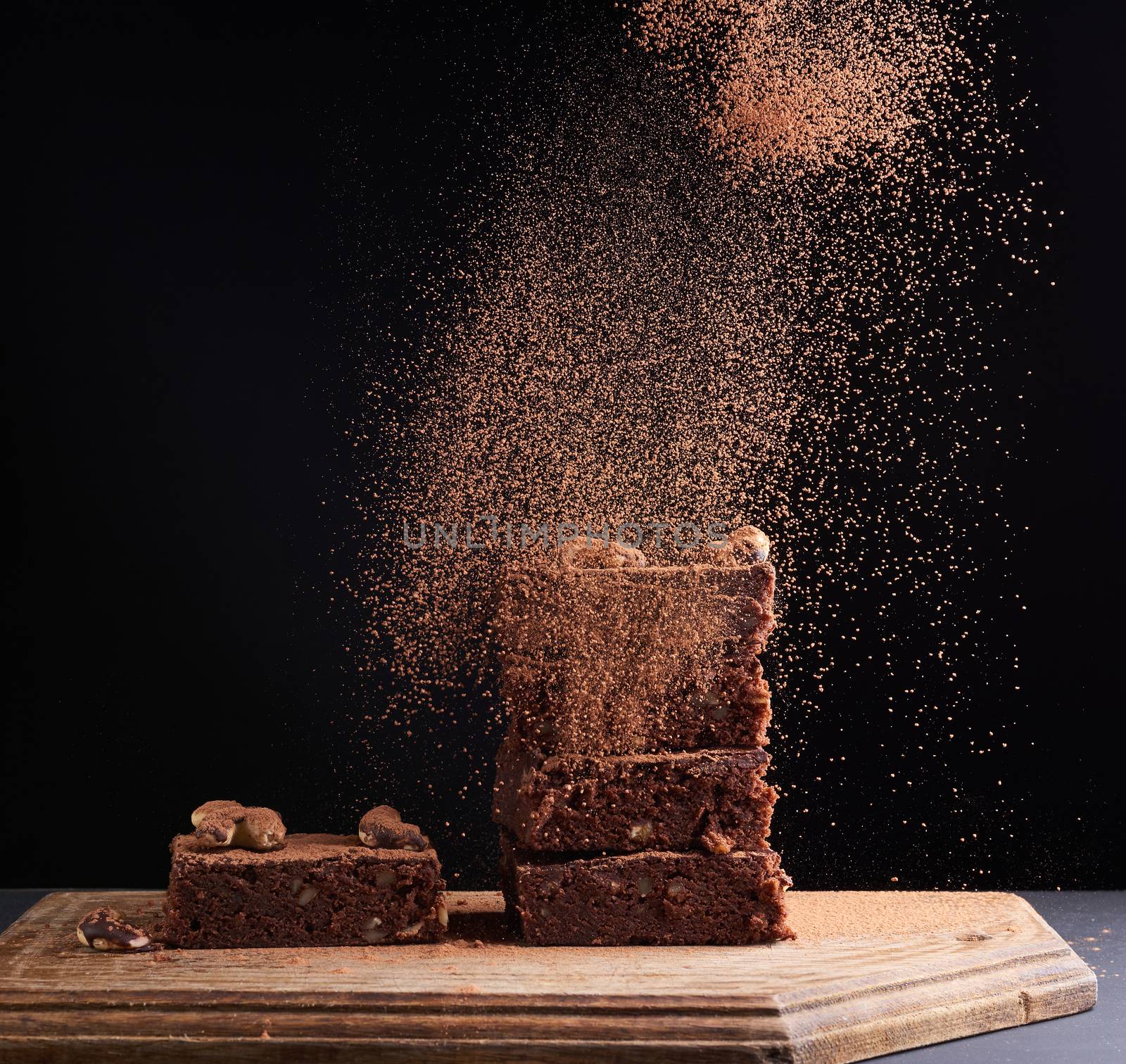 stack of square baked brownie pieces sprinkled with cocoa powder, particles froze in the air against a dark background,  delicious chocolate dessert