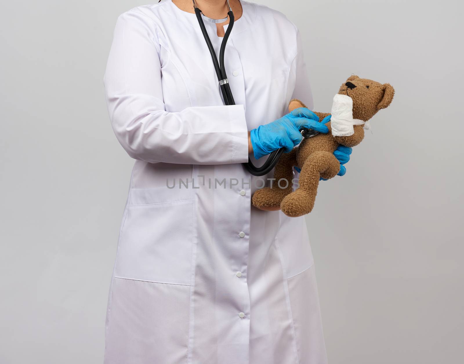female medic holds brown teddy bear with paw bandaged in white bandage and listens to toy with stethoscope, concept of pediatrics and animal treatment