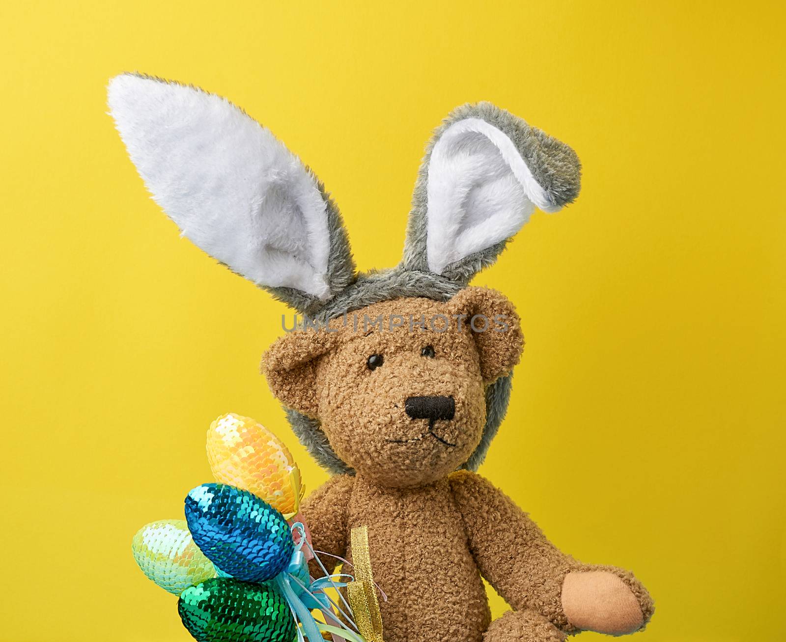 cute brown teddy bear holding colorful Easter eggs, wearing a rabbit mask with long ears on his head, funny holiday card