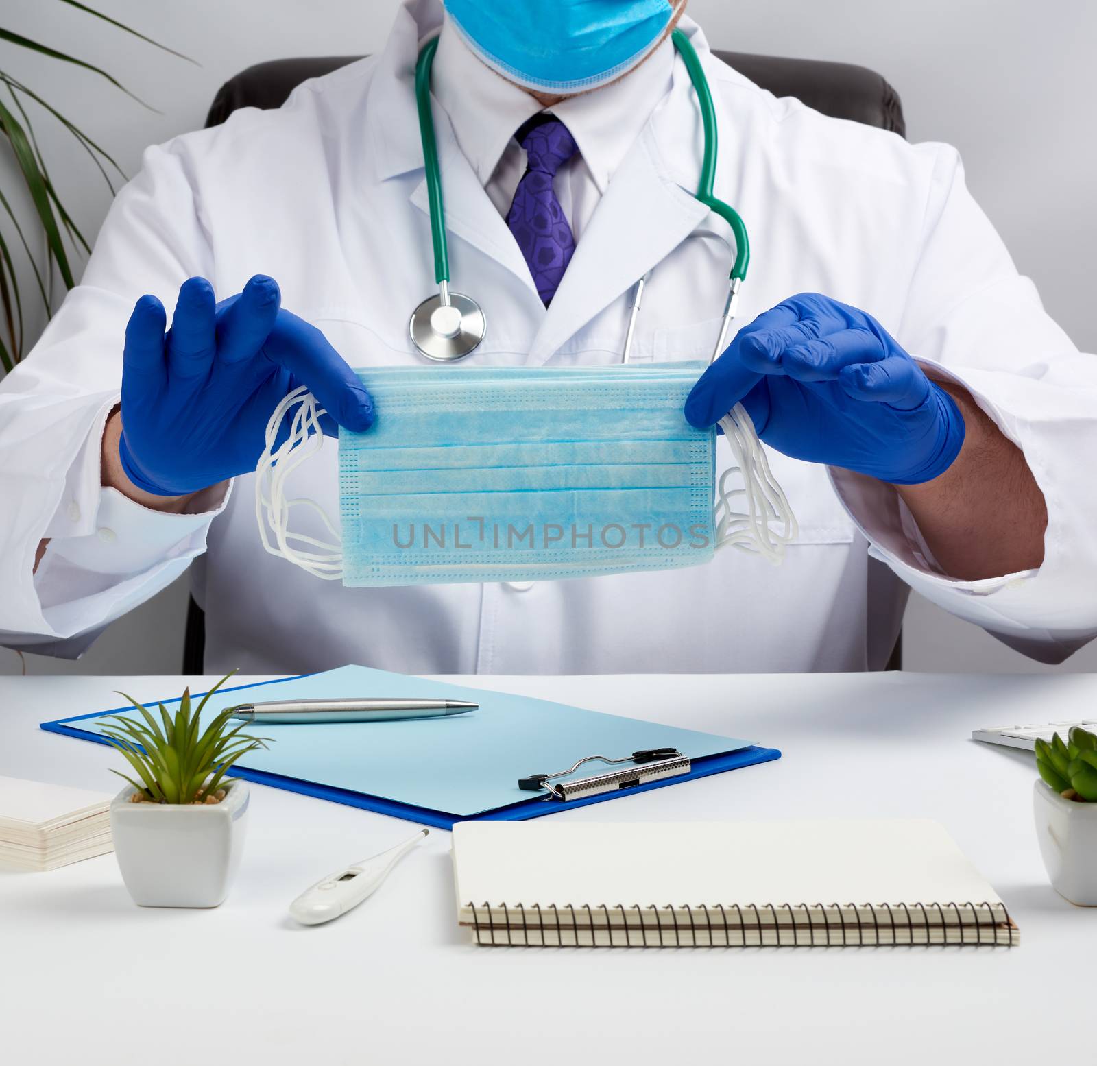male doctor in a white coat and blue latex gloves sits at a white desk and shows a stack of disposable medical masks, protective agent against viruses and stopping the spread of the epidemic and pandemic