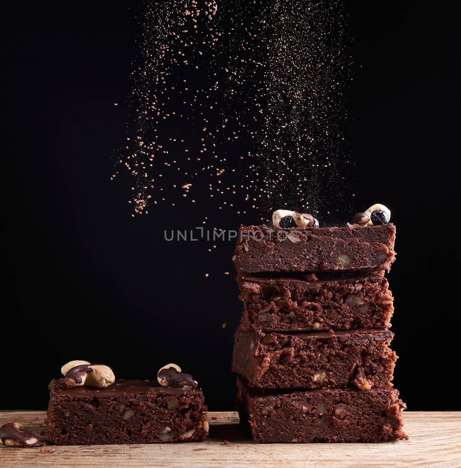 stack of square baked brownie pieces sprinkled with cocoa powder, particles froze in the air against a dark background,  delicious and delicious chocolate dessert