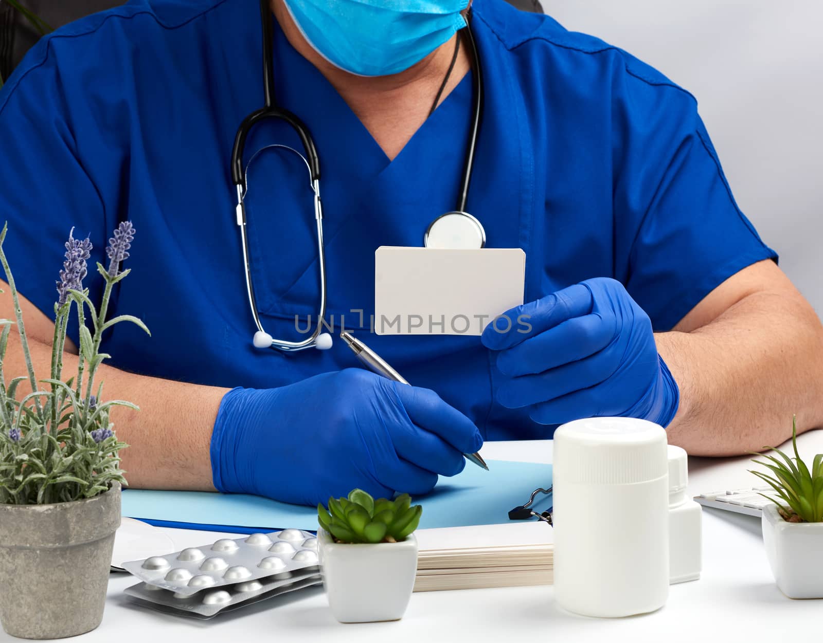 male doctor in a blue uniform sits at a white table and writes in a paper notebook, wearing sterile gloves on his hands, concept of receiving patients in the office