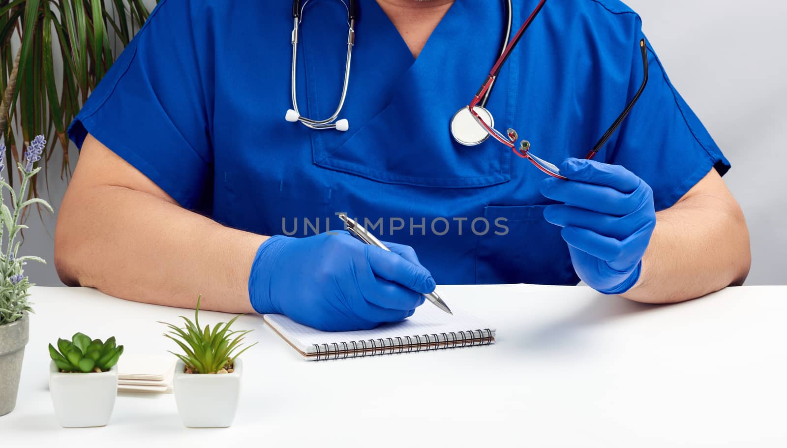 a male doctor in a blue uniform sits at a white table and writes in a paper notebook, wearing sterile gloves on his hands, concept of receiving patients in the office