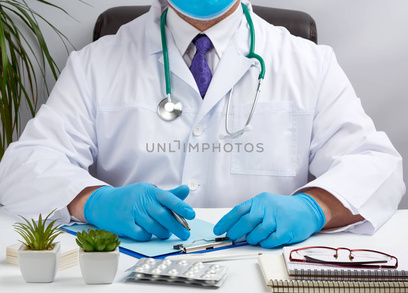 doctor in a white uniform sits at a white table and writes in a paper notebook, wearing sterile gloves on his hands, concept of receiving patients in the office