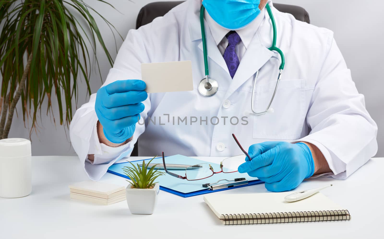 doctor in a white medical coat sits at a table in a brown leather chair and holds a rectangular white blank business card in his hands, medical office