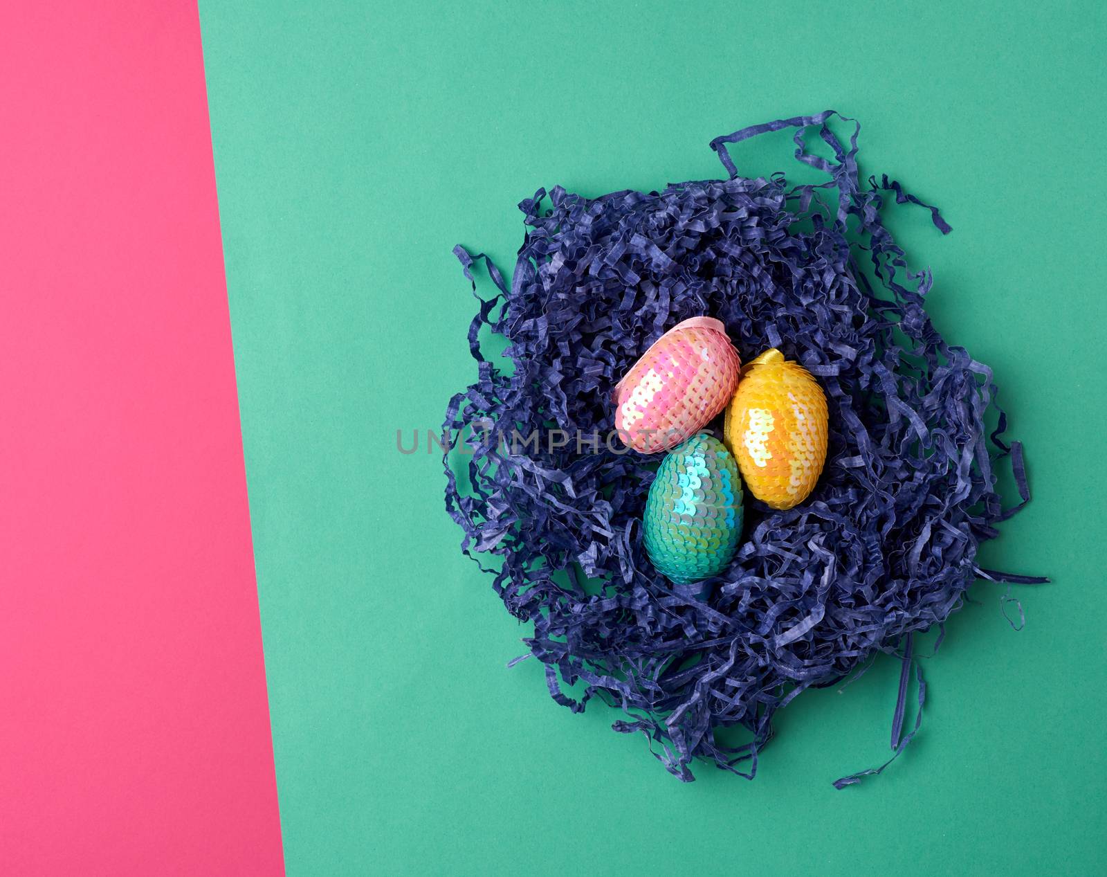 decorative Easter eggs from sequins lie in a nest of strips of paper on a colored background, top view