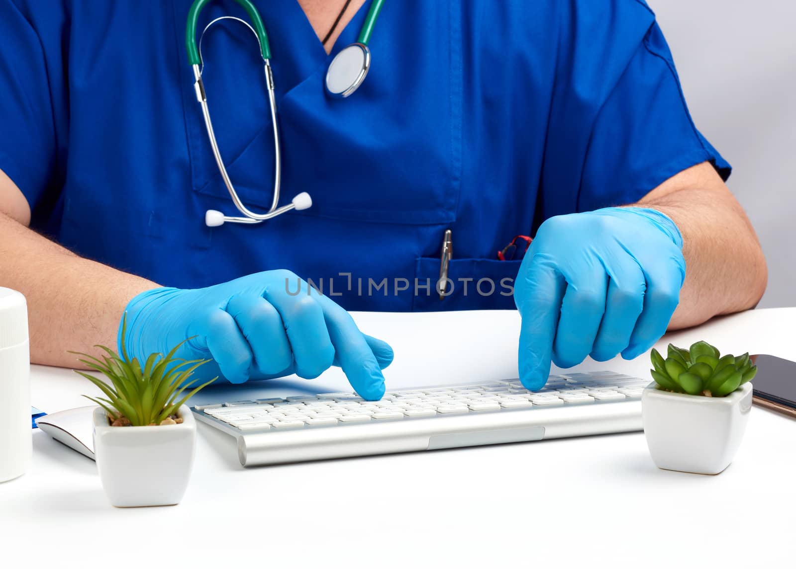 male doctor sitting at a white table in blue uniform and latex gloves, specialist typing on a keyboard, medical office for receiving patients, close up