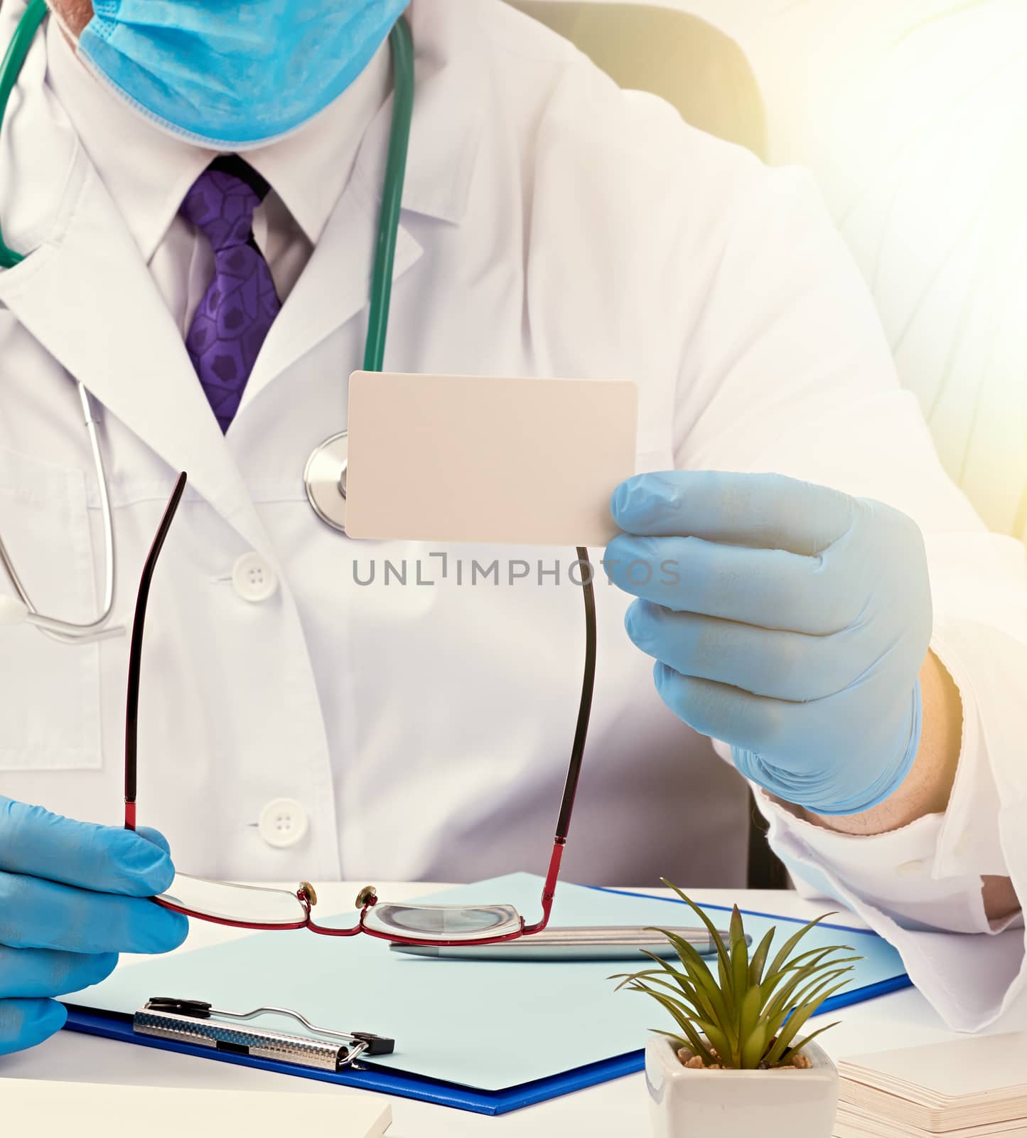 doctor in a white medical coat sits at a table in a brown leather chair and holds a rectangular white blank business card in his hands, medical office