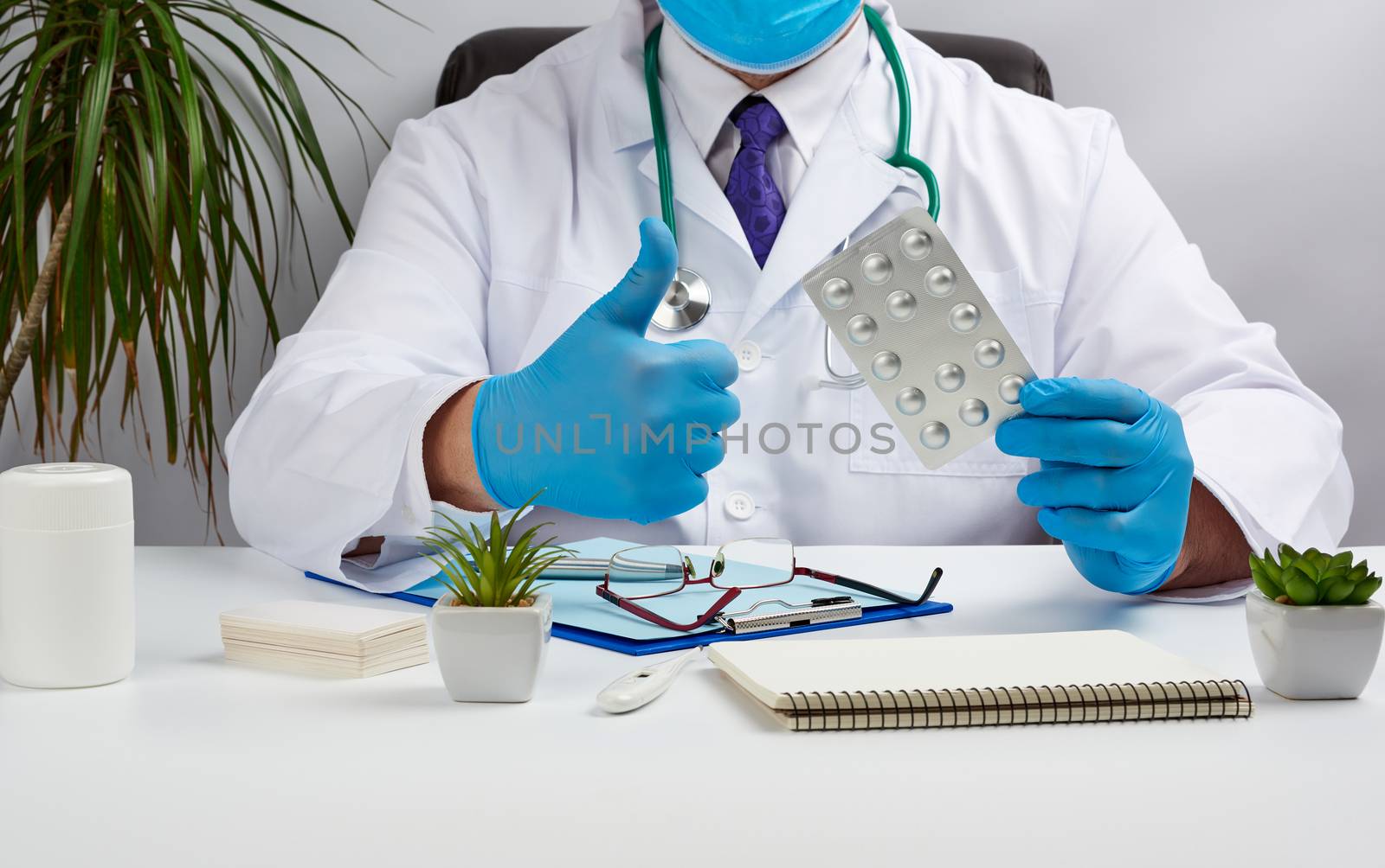 male doctor in a white coat and blue latex gloves sits at a white desk in his office and shows a pack of pills in a blister, with his other hand shows a gesture like